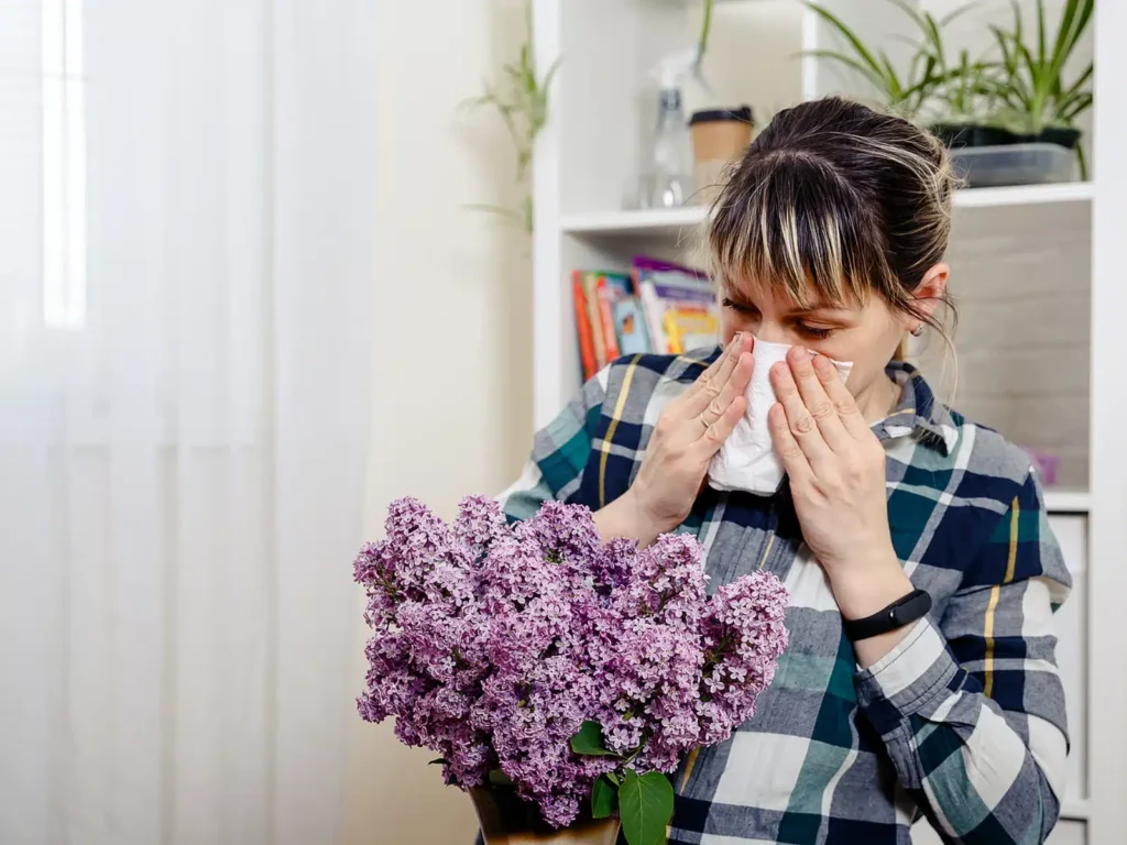 Woman sneezing into a tissue while holding a bouquet of flowers, representing common fall allergy symptoms and triggers.